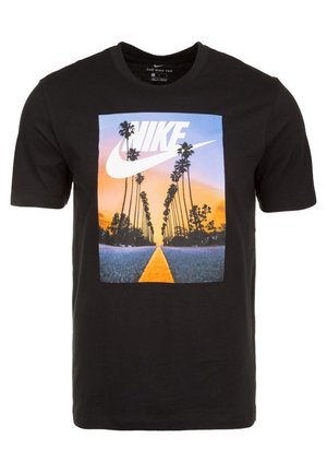 T-shirt noir Nike avec une scène de route au coucher du soleil, comprenant de grands palmiers et un grand logo blanc Nike dans le ciel.
