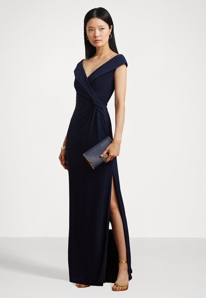 Abito da sera blu navy con design avvolgente, maniche scoperte sulle spalle e uno spacco alto fino della coscia. Abbinato a una clutch scura e tacchi dorati con cinturini.