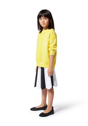 Sweatshirt jaune avec des poignets côtelés, jupe blanche à bords festonnés avec des rayures noires, et ballerines noires à pois blancs. Design simple et moderne.