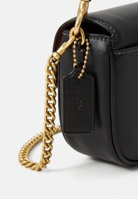 Black leather bag corner with gold chain strap and attached black Coach New York tag on white background.