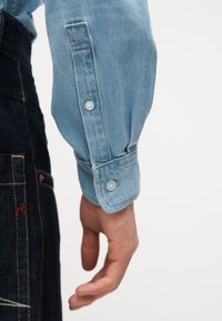 Κοντινό ενός χεριού ατόμου που φοράει ανοιχτό μπλε πουκάμισο από τζιν με μανίκια μακριά και κουμπωτά μανσέτα, να ξεκουράζεται δίπλα σε σκουρόχρωμο τζιν παντελόνι.