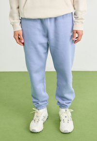 Pantaloni de trening bleu deschis dintr-o țesătură moale, cu manșete elastice și buzunare laterale. Asortați cu pantofi sport albi.