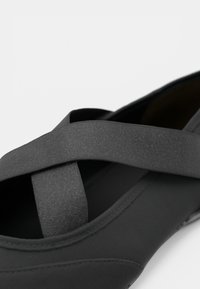 Sandal preto fechado com tiras elásticas largas em forma de cruz e textura de tecido suave, contra um fundo branco liso.