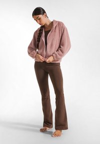 Fluffy roze zip-up jasje met een capuchon, gecombineerd met strakke bruine flare broek. Zachte texturen, losse mouwen, en een casual ontwerp.