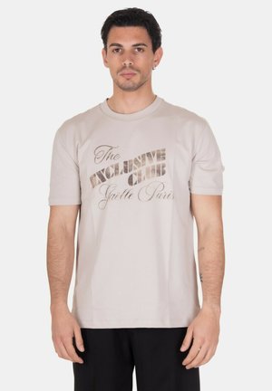 Uomo che indossa una T-shirt beige a maniche corte con la scritta "The Exclusive Club Gaëlle Paris", abbinata a pantaloni neri, in piedi su sfondo neutro.