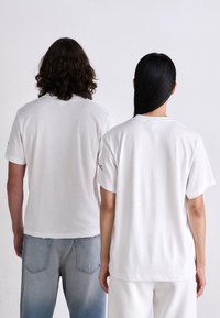Dos personas están de pie de espaldas, vistiendo camisetas blancas sencillas; una con jeans de un azul claro y la otra con pantalones blancos, frente a un fondo liso.