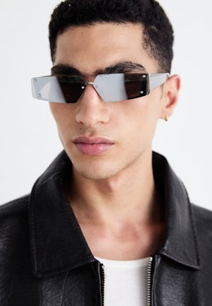 Gafas de sol angulares de tonalidad gris con detalles en plata, sobre un fondo claro. Las lentes presentan una forma geométrica única.