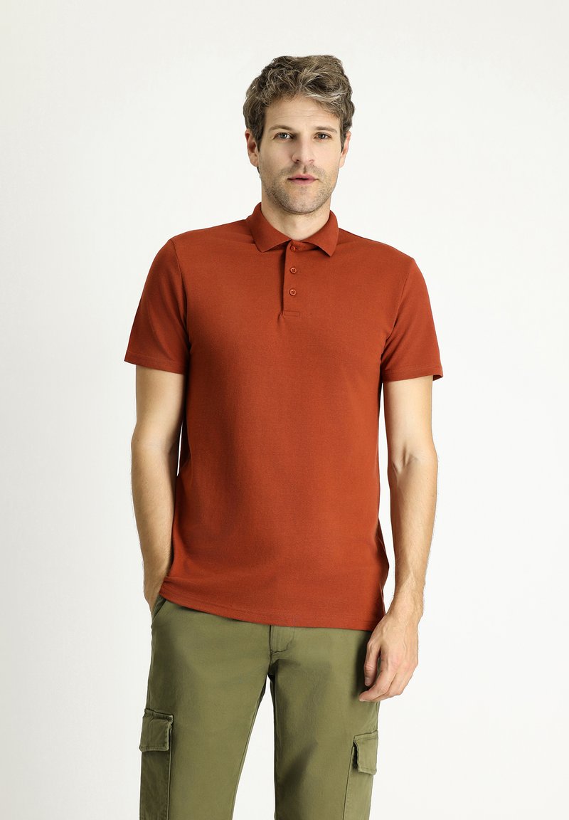 Camisa polo de color óxido con un collar clásico, tapeta de tres botones y mangas cortas. Llevada con pantalones cargo oliva, que tienen bolsillos laterales.
