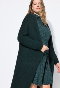 Vrouw in een donkergroene fluffy cardigan over een geknoopte jurk met panterprint, poseert tegen een effen witte achtergrond.