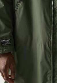 Donkere hand naast de mouw van een olijfgroene jas met een patch waarop "HYDRAFORT ACTIVE PERFORMANCE" staat en een zichtbare rits aan de voorkant.