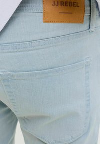 Jean en denim bleu clair montrant la poche arrière et l’étiquette de marque en cuir marron « JJ REBEL » sur la ceinture.