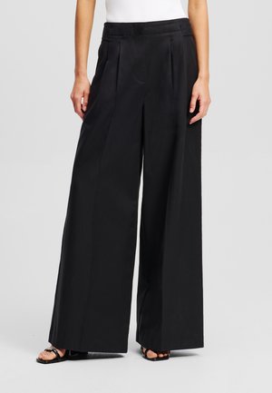 Zwarte wijdvallende broek van soepel stof, met plooien en een hoge taille. Gemaakt met een nauwsluitend wit topje en zwarte hakken.