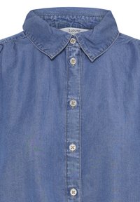 Modrá chambray košeľa so špicatým golierom a piatimi krémovými gombíkmi vpredu, s viditeľným prešitím pozdĺž okrajov a švov.