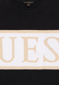 Čierny sveter s širokým bielym pásom a vyvýšeným zlatým nápisom "GUESS" cez hruď. Okrúhly výstrih a jemná textúra.