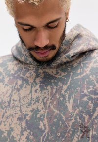 Hætte sweatshirt i et camo mønster af beige, grøn og brun. Blødt stof med en subtil tekstur og et lille broderet logo på brystet.