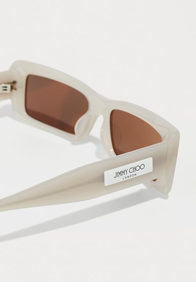 Lunettes de soleil couleur crème avec des verres marron, monture rectangulaire et étiquette "Jimmy Choo London" sur la branche.