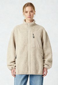 Giacca in pile beige con collo alto, cerniera frontale e tasca sul petto. Realizzata in materiale morbido e testurizzato, dotata di due tasche laterali.