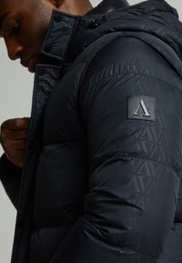 Chaqueta de plumas azul marino con un diseño acolchado, que presenta un parche con logo en la manga, un tejido texturizado y un cuello alto con cierres a presión.