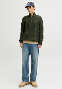 Un pull en tricot vert foncé avec un demi-zip, texture côtelée large, associé à un jean baggy bleu clair et des chaussures beiges. Une casquette de baseball bleue complète le look.