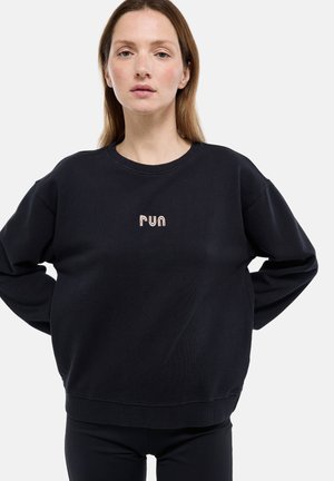 Femme portant un sweat-shirt noir avec le texte « run » en rose clair, debout les mains sur les hanches devant un fond blanc uni.