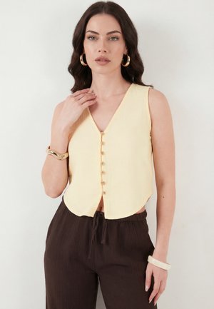 Sötét hajú nő ujjatlan halvány sárga gombos felsőt, sötétbarna nadrágot, aranykarika fülbevalót és karkötőket visel egyszínű háttér előtt.