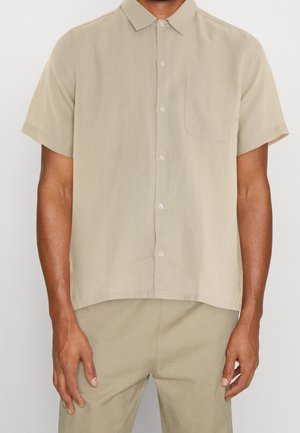 Mand iført beige kortærmet skjorte med knapper og brystlomme samt matchende beige bukser står mod en ensfarvet baggrund.
