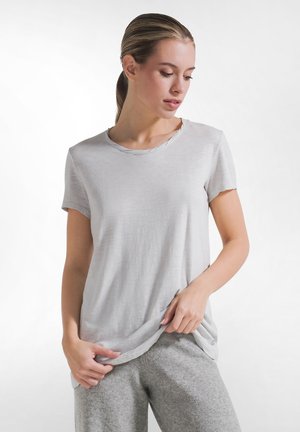 Vrouw met het haar naar achter gebonden, die een lichtgrijs T-shirt met korte mouwen en een grijze broek draagt, kijkt omlaag en houdt de zoom van haar shirt vast.