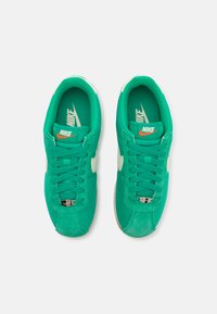 Ténis verdes com um cabedal liso e detalhes em camurça, apresentando o swoosh branco da Nike, atacadores e um interior almofadado. Solado de borracha visível.