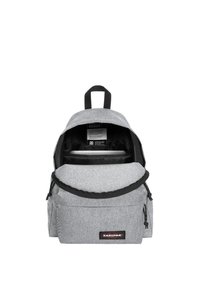 Mochila de tela gris con parte superior redondeada, compartimento principal con cremallera, bolsillo frontal y correas y acentos negros. Etiqueta visible en el interior.