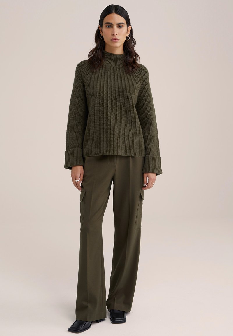 Zielona oliwkowa swetrowa bluzka z prążkowanego materiału, z wysokim kołnierzem i szerokimi mankietami, zestawiona z oliwkowymi spodniami cargo o szerokich nogawkach z bocznymi kieszeniami oraz czarnymi butami.