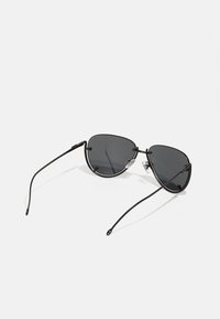 Lunettes de soleil noires en métal avec des verres gris, forme œil de chat, design minimaliste, branches fines et accents matériels subtils au niveau des charnières.