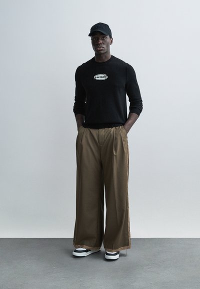 Black knit sweater with logo, tan wide-leg trousers, and black cap. Black sneakers with white accents. Smooth textures, casual fit.