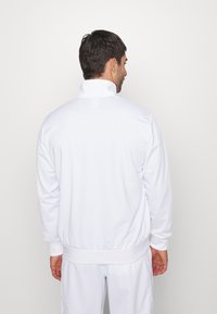 Chaqueta de chándal blanca con cremallera y un alto cuello, puños y cintura elásticos. Textura suave, diseño minimalista, sin patrones ni acentos visibles.