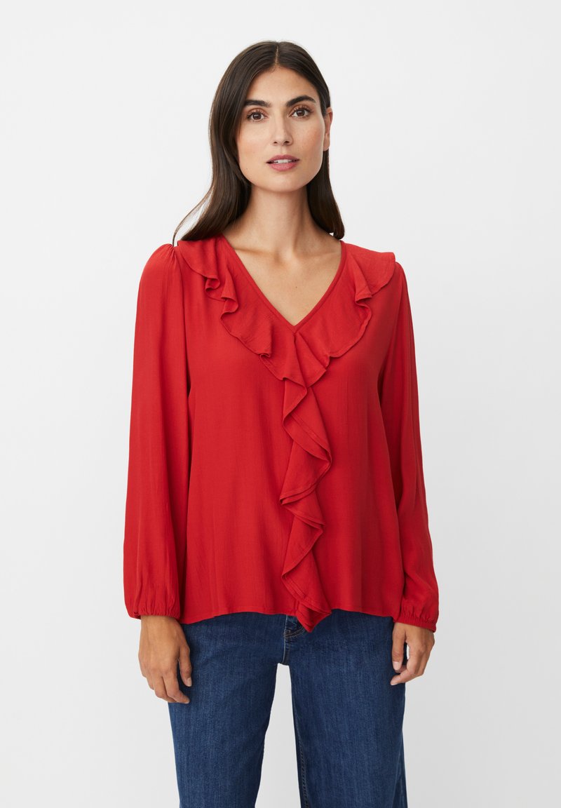 Rote Bluse mit V-Ausschnitt, die entlang der Vorderseite Rüschendetails und lange, leicht geraffte Ärmel hat. Getragen mit blauen Jeans.
