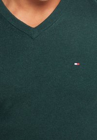 Close-up van een groene V-hals trui met een klein geborduurd rechthoekig logo in rood, wit en blauw op de borst.
