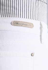 Jean en denim blanc avec une patch en cuir beige sur la taille, présentant des coutures standard et une poche avant avec un rivet argenté.