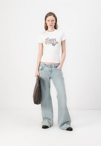 Witte katoenen t-shirt met bloemenprint, gecombineerd met lichtblauwe wijde jeans. Model houdt een bruine tas vast en staat tegen een effen achtergrond.