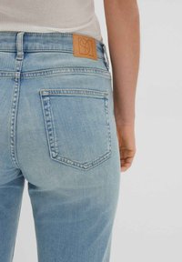 Light blue jeans feature a mid-rise waist, classic five-pocket design, and a leather patch with a logo at the back. Faded texture visible.