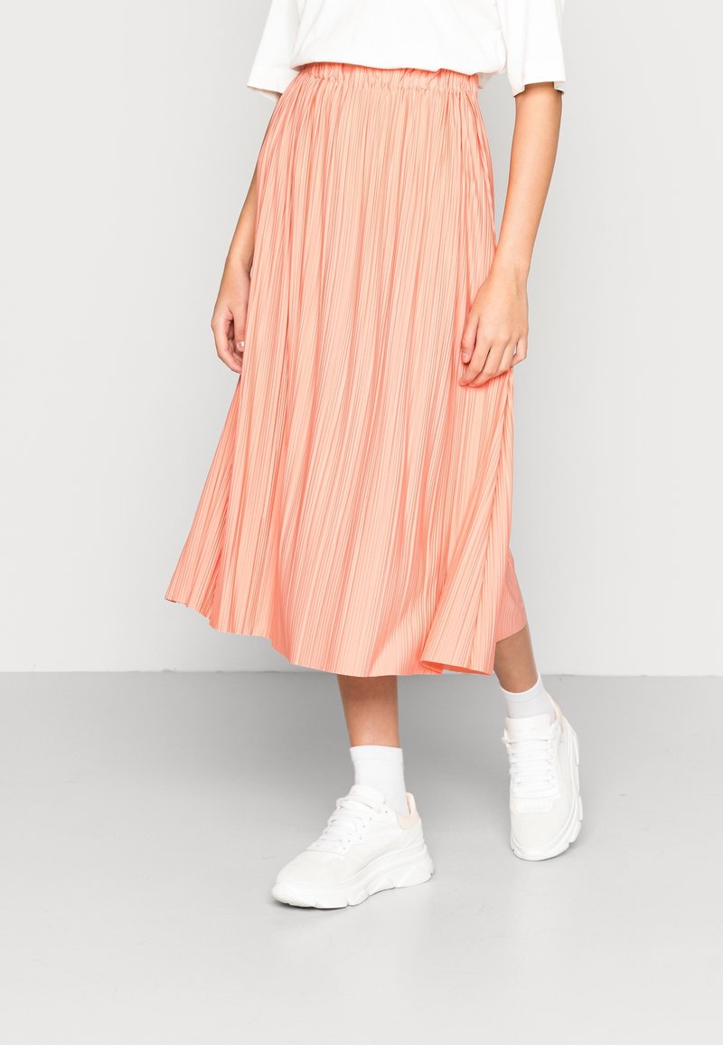 Femme portant une jupe midi plissée corail, un haut blanc rentré, des chaussettes blanches et des baskets blanches épaisses, debout sur un sol gris clair.