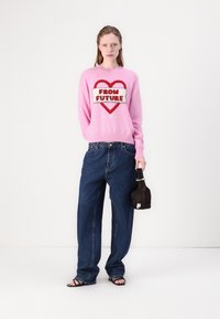 Maglione rosa lavorato a maglia con la scritta "DAL FUTURO" in rosso. Jeans in denim blu larghi e borsa nera. Modella che indossa sandali neri.