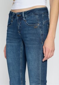 Slim fit džíny v tmavě modrém džínovém materiálu, které mají pět kapes, obroušené švy a stříbrný knoflík na zapínání s logem.