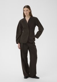 Sieviete stāv, valkājot tumši brūnu, vaļēju kostīmu bez apkakles žaketi un platajiem bikšu galiem, pozējot ar vienu roku kabatā.