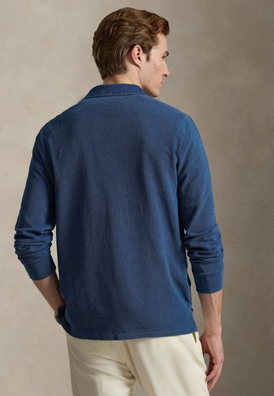 Navy blue long-sleeve polo shirt with a textured knit. Features a buttoned side and classic collar. Worn with light-colored trousers.