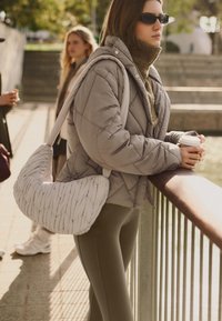 Pikowana szara kurtka z wysokim kołnierzem, zielony dziergany sweter, dopasowane oliwkowe legginsy oraz teksturowana biała torebka. Trzyma kubek z kawą.
