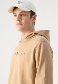Felpa beige realizzata in tessuto morbido con tasca a marsupio, con logo grigio e piccolo emblema a righe. Cappello bianco con testo ricamato.