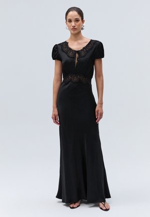 Femme portant une longue robe noire avec des détails en dentelle sur le décolleté et la taille, debout devant un fond uni gris clair.