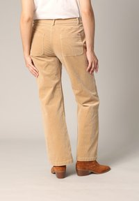 Pantalons large en velours côtelé beige avec deux poches arrière, tissu texturé et ourlet net, associés à des bottes à talons marron.