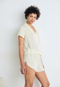 Ensemble de pyjama jaune clair à manches courtes comprenant une chemise à boutons et un short assorti à l'ourlet arrondi, fabriqué dans un tissu doux et transparent.