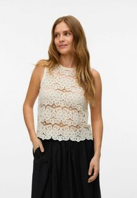 Krajková tílko v off-white barvě s květinovým vzorem, vysokým výstřihem a bezrukávovým designem. Spárováno s černou, objemnou sukní.