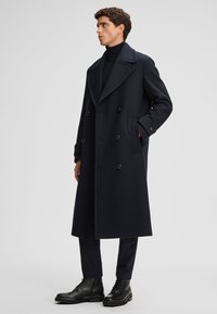 Marineblauer doppelt genieteter Überzieher mit breiten Revers, seitlichen Taschen und schwarzen Knöpfen; getragen über einem schwarzen Rollkragenpullover und schmal geschnittenen Hosen.